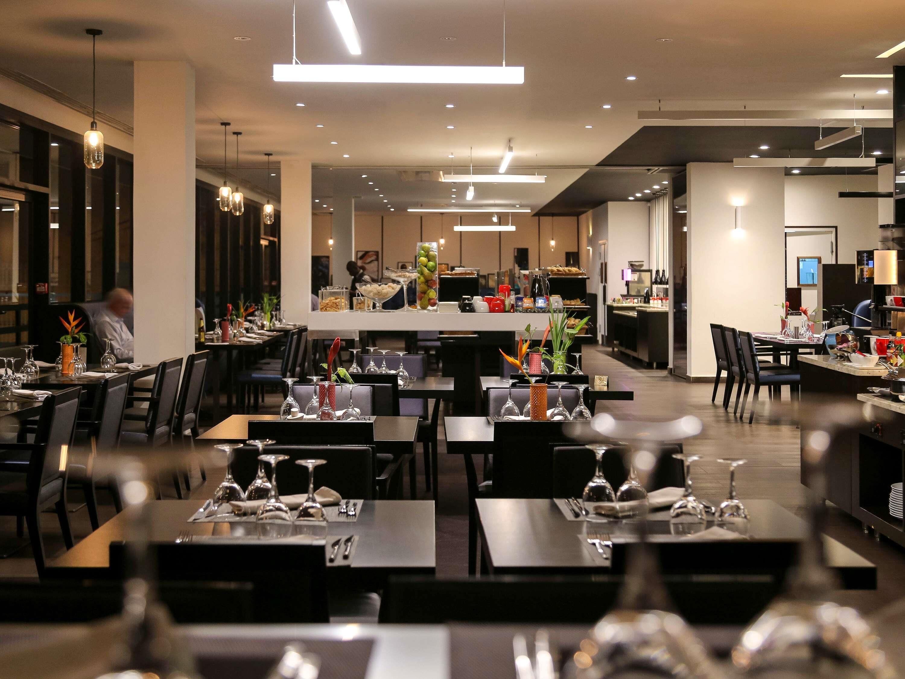 Un restaurante moderno con varias mesas, iluminación colgante y una cocina a la vista.