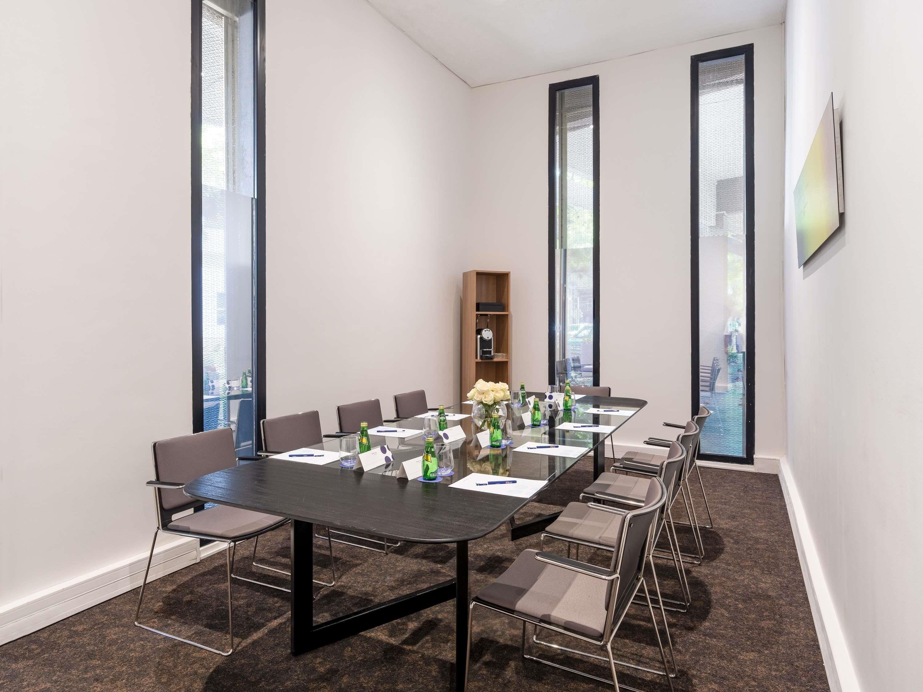 Un salón de conferencias con una larga mesa, sillas, botellas de agua y un reloj en la pared.