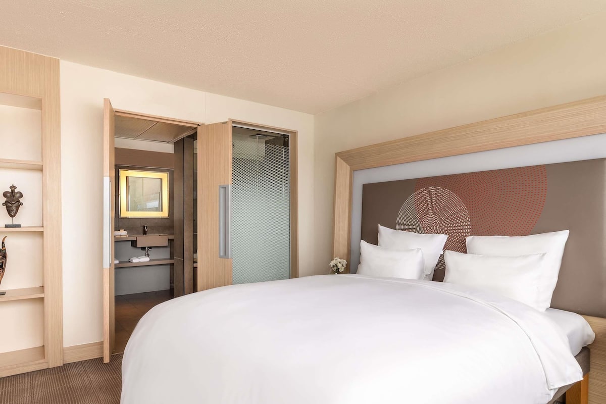 A hotel room with a large bed, a wooden headboard, a bathroom with a mirror, and a small table with a vase.