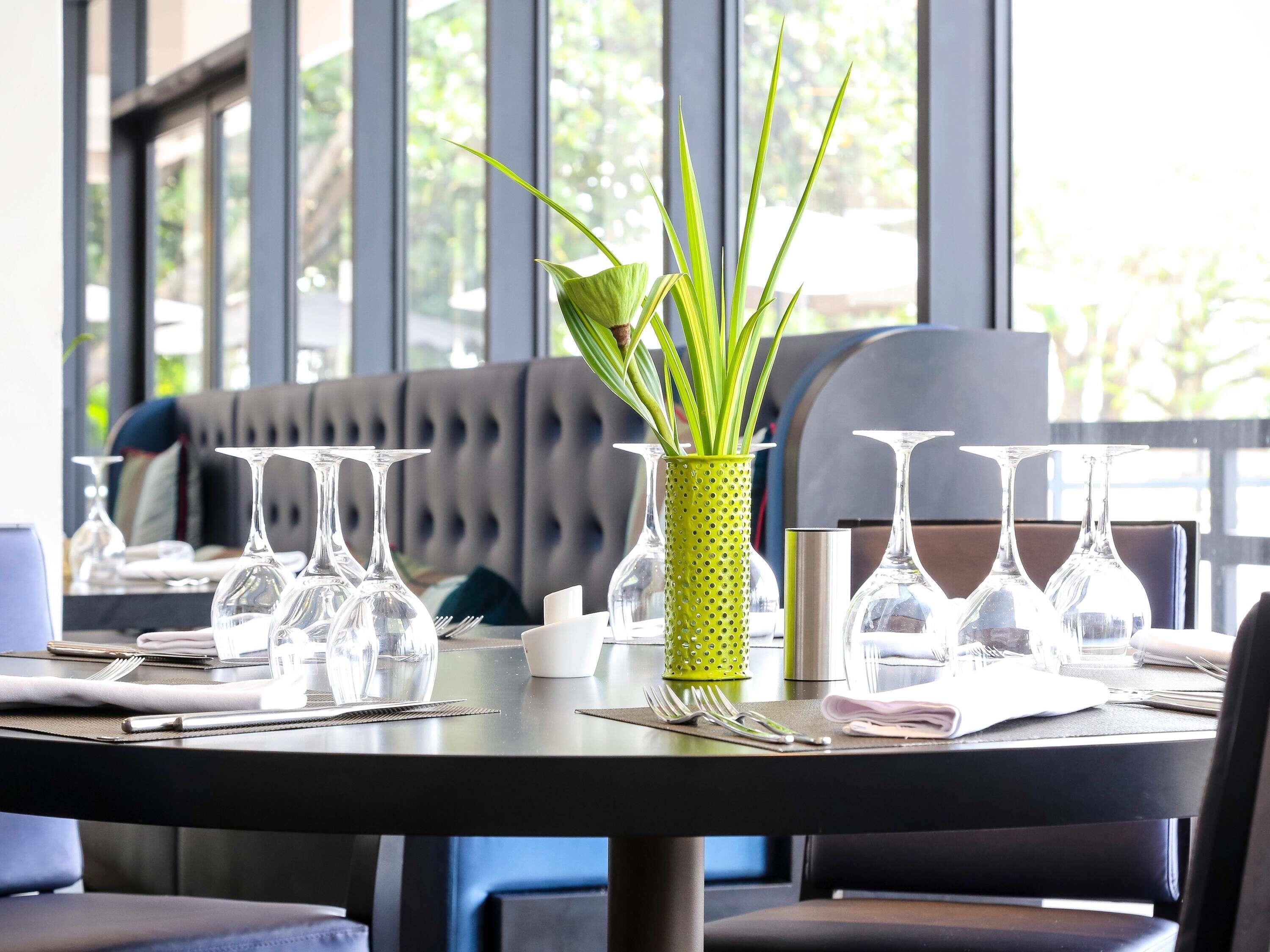 A dining table set with clear glassware, a green plant in a textured pot, and a grey upholstered bench in the background.