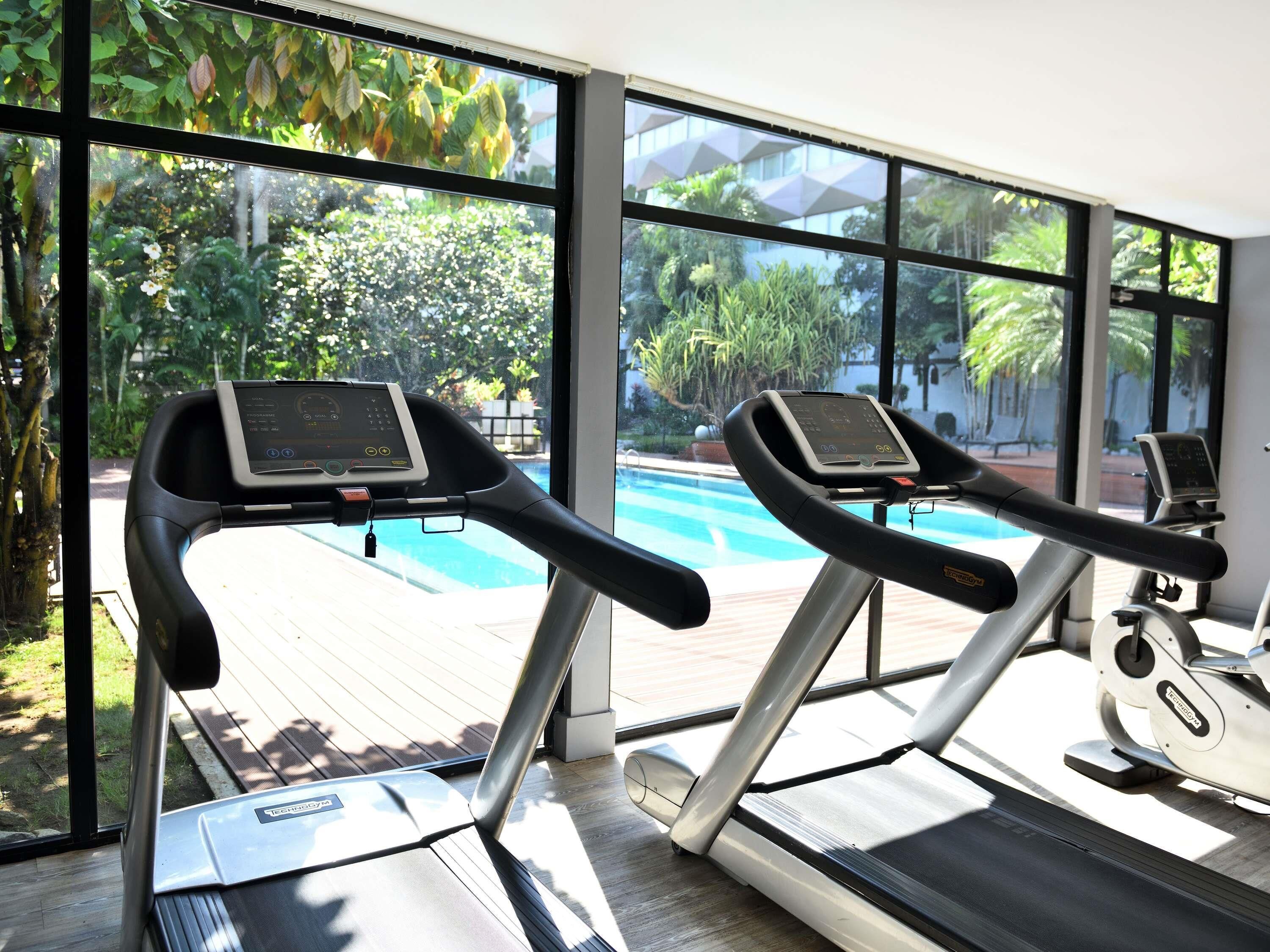 Dos cintas de correr con pantallas digitales, ubicadas en un gimnasio con amplias ventanas que dan a una piscina y exuberante vegetación.