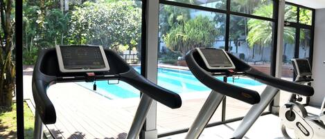 Dos cintas de correr con pantallas digitales, ubicadas en un gimnasio con amplias ventanas que dan a una piscina y exuberante vegetación.