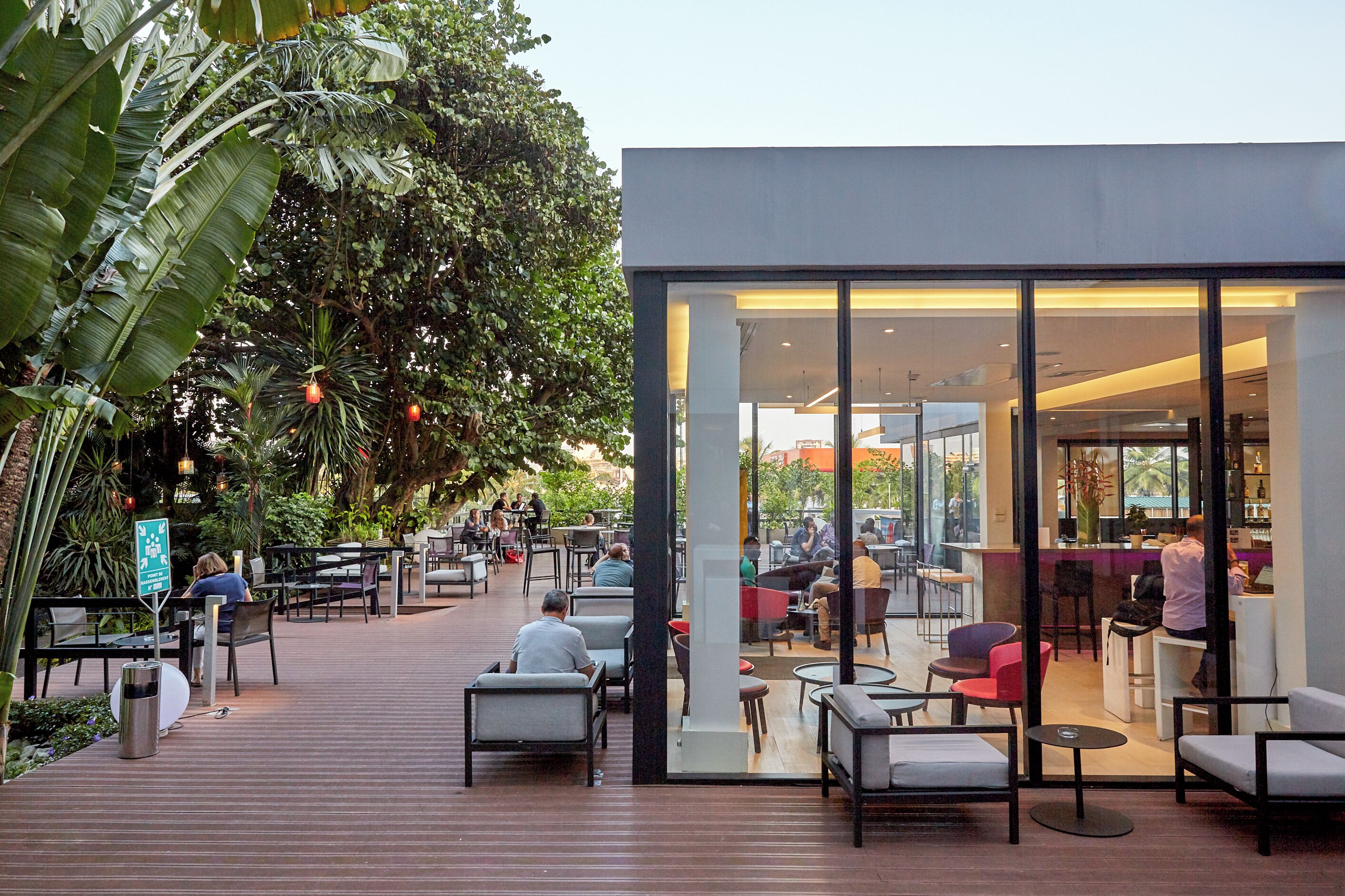 An outdoor café area with wooden decking, seating, and a glass wall leading to an interior space.