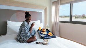 Una habitación de hotel con una cama, una bandeja con desayuno, una ventana con vistas a edificios y un cabecero estampado.