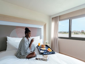 A hotel room with a bed, a tray with breakfast, a window with a view of buildings, and a patterned headboard.