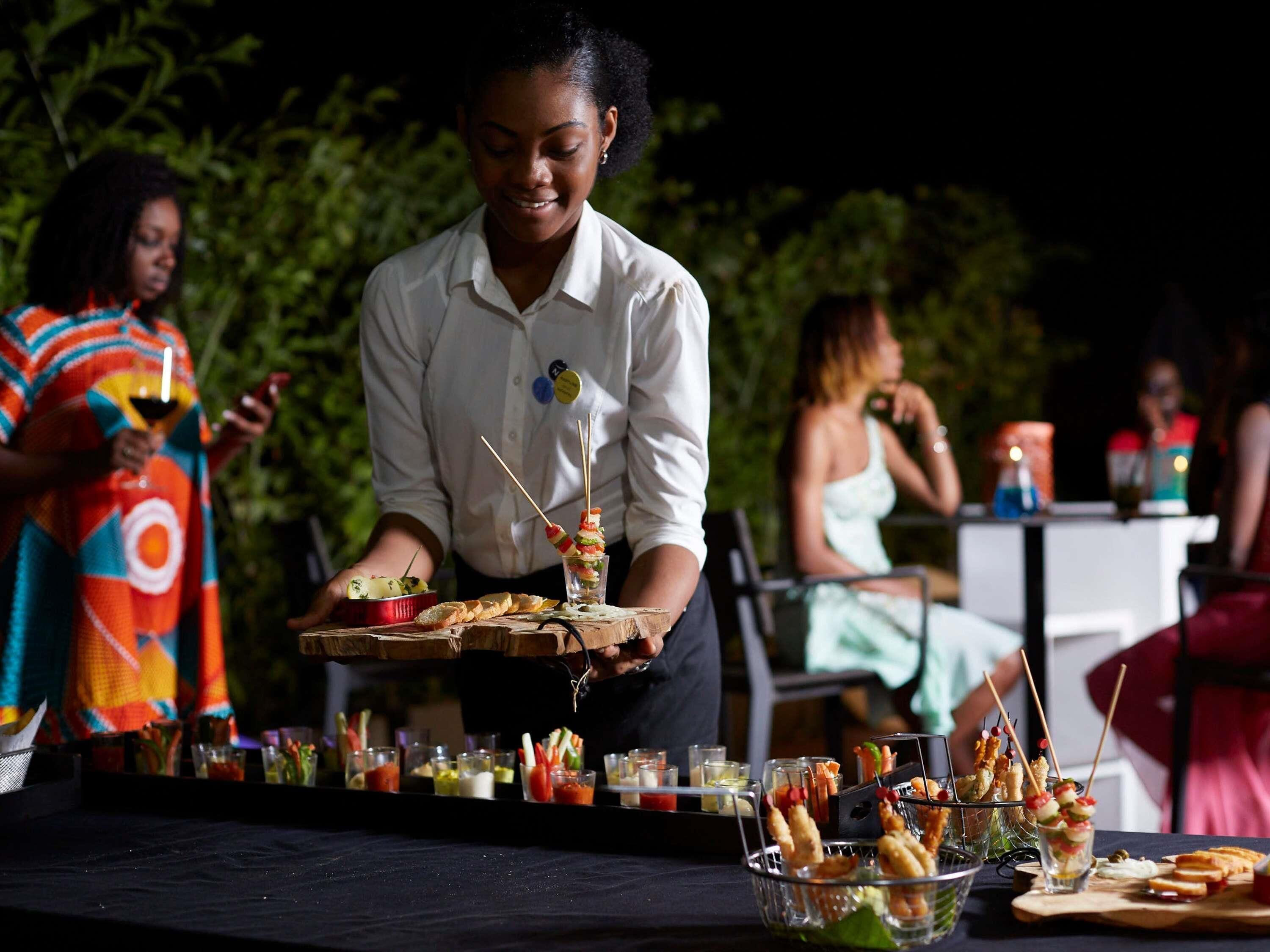 Una persona sirviendo comida en un evento al aire libre, con varios platos y bebidas sobre una mesa.