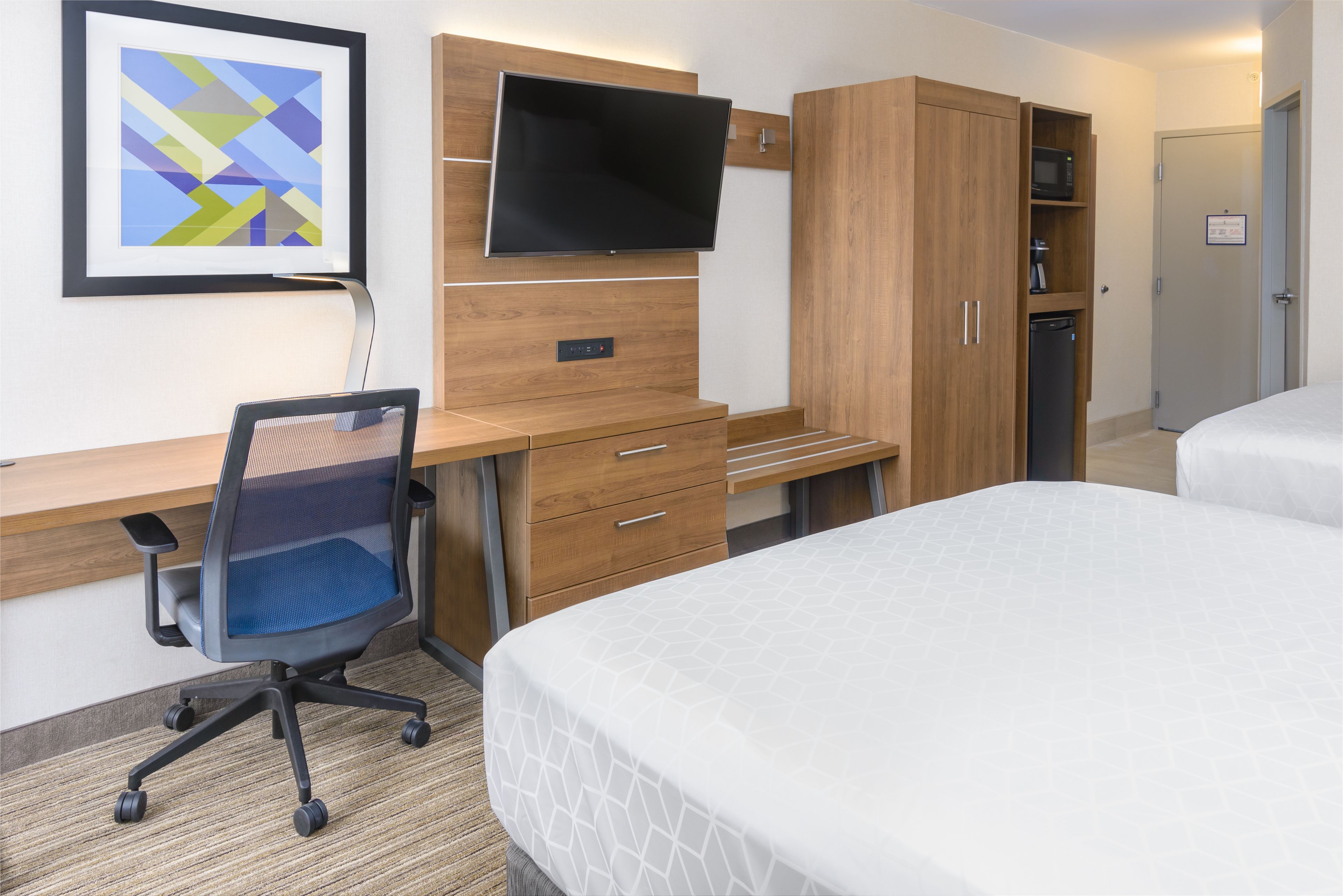 Habitación de hotel con cama, escritorio con silla, televisor montado en la pared y armario.