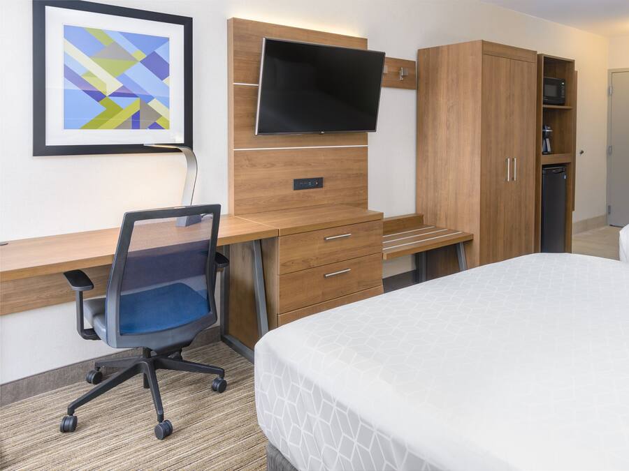 A hotel room with a bed, a desk with a chair, a TV mounted on the wall, and a wardrobe.