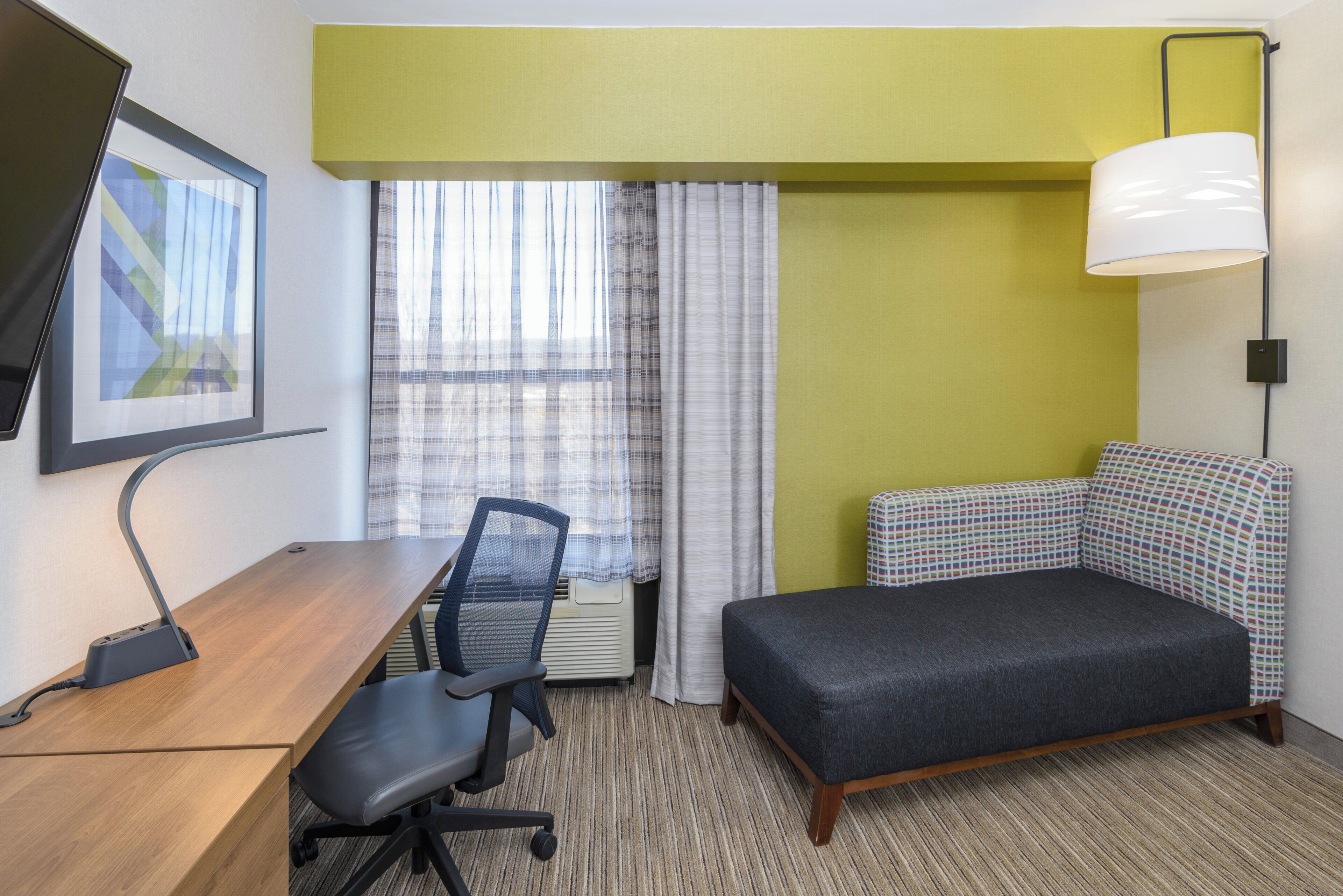 A hotel room with a desk, chair, bed, and a large window with curtains.