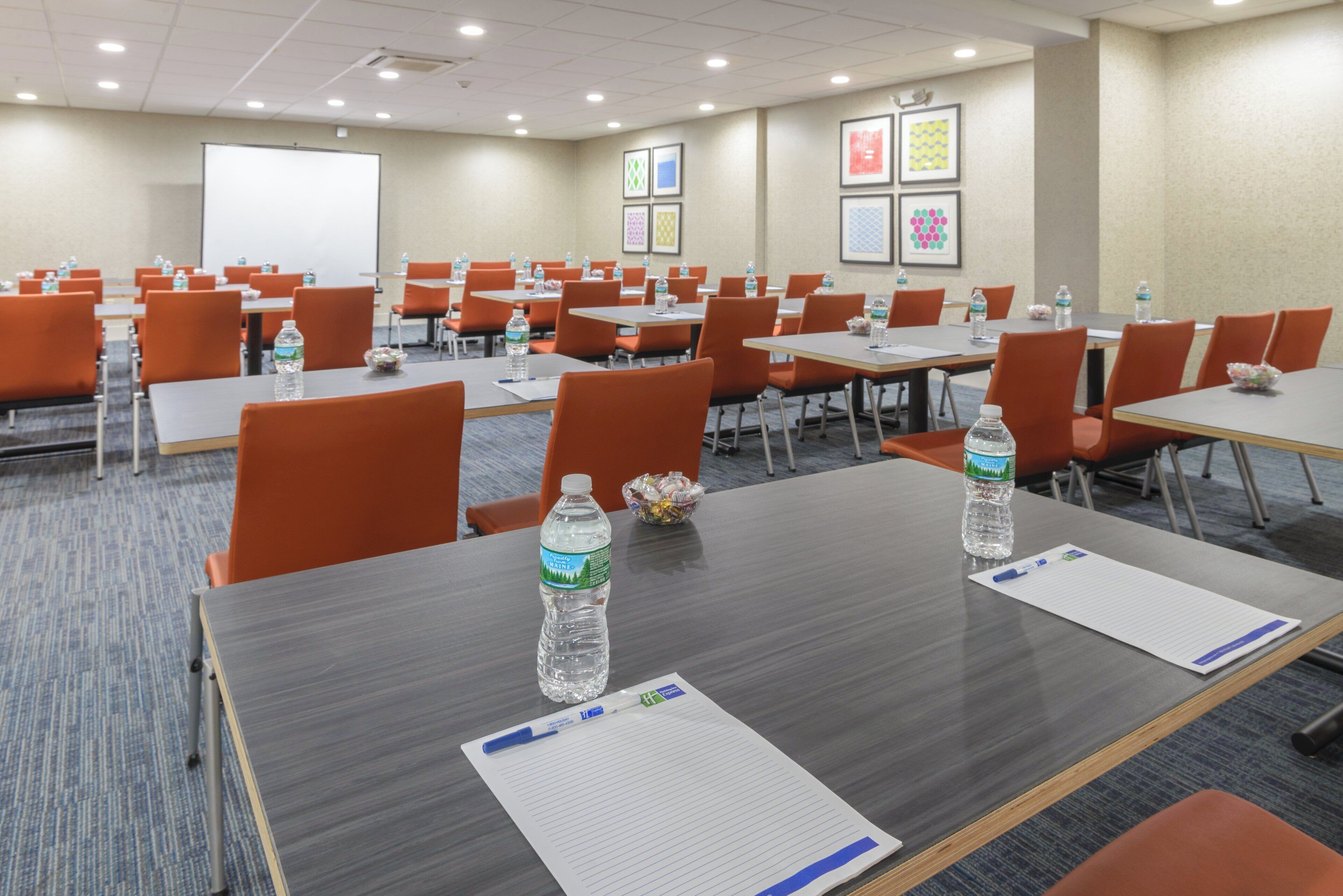 Une salle de conférence équipée de tables, de chaises, de bouteilles d’eau et d’écrans de présentation.