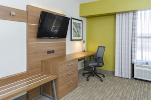 A hotel room with a wooden desk, a chair, a flat-screen TV, a framed picture, and a window with curtains.