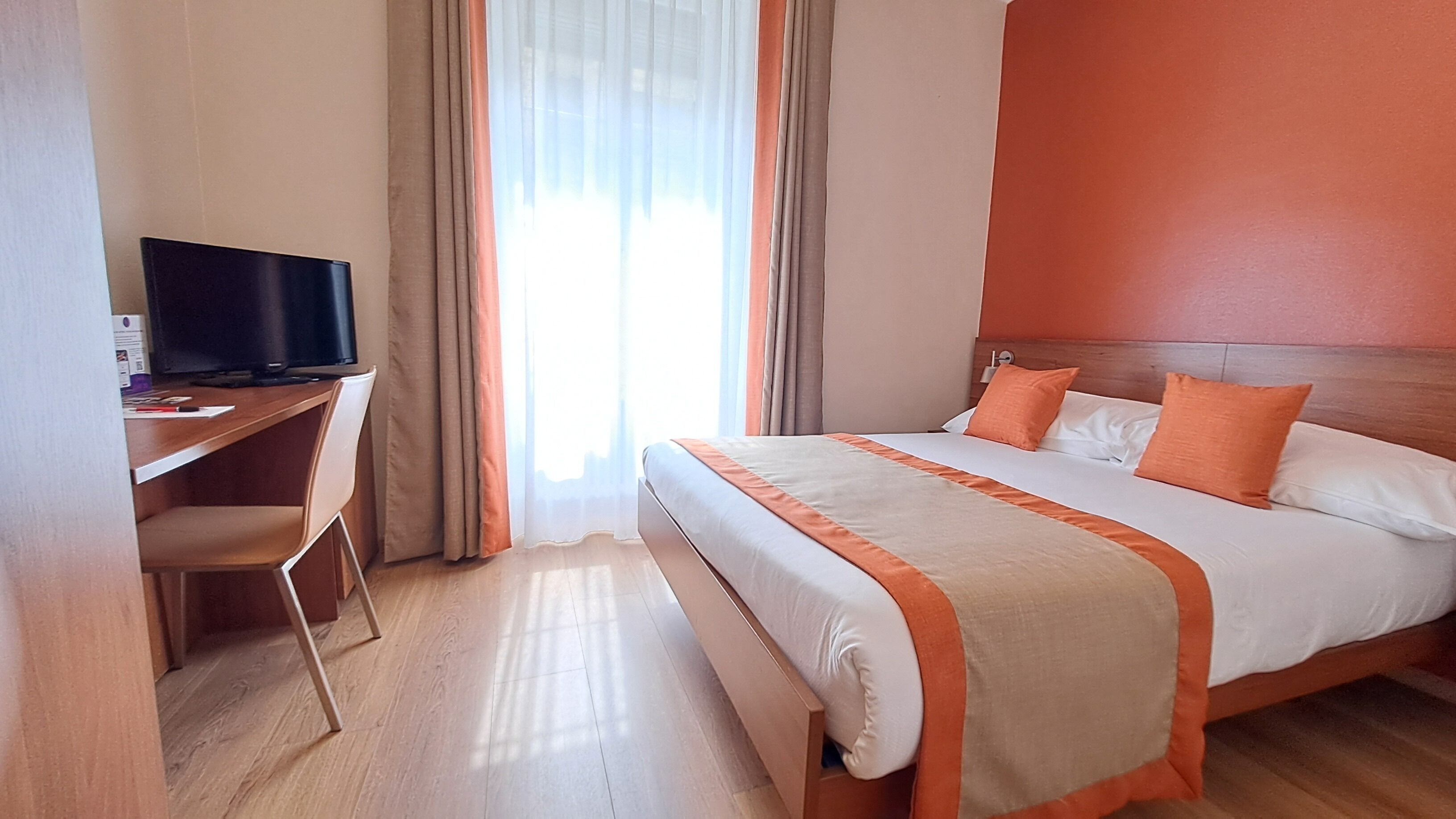 A hotel room with a bed, a desk with a computer, and a window with curtains.