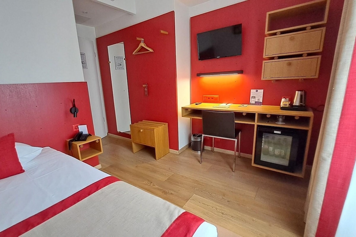Una habitación de hotel compacta con cama, escritorio, kitchenette pequeño y televisión.