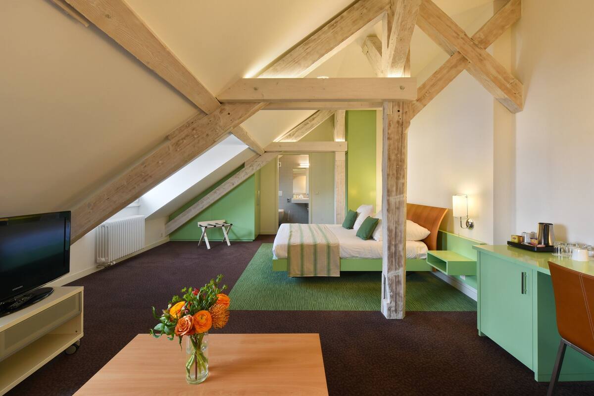 A hotel room with a lofted ceiling, a bed, a desk, a TV, and a small table with a vase of flowers.