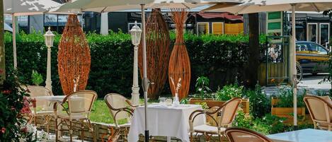 An outdoor dining area with wicker chairs and white tables, covered by striped canopies.