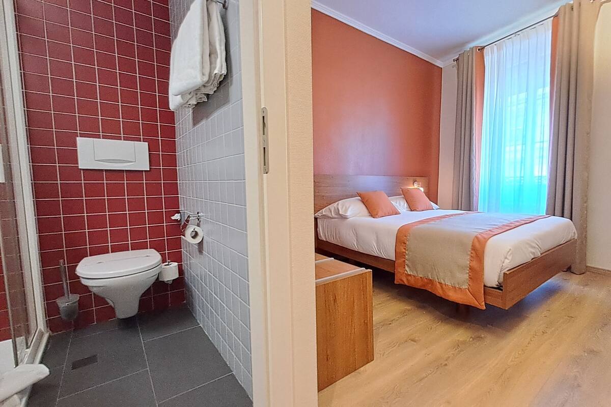 A hotel room with a bed, a desk, a chair, a toilet, and a window with curtains.
