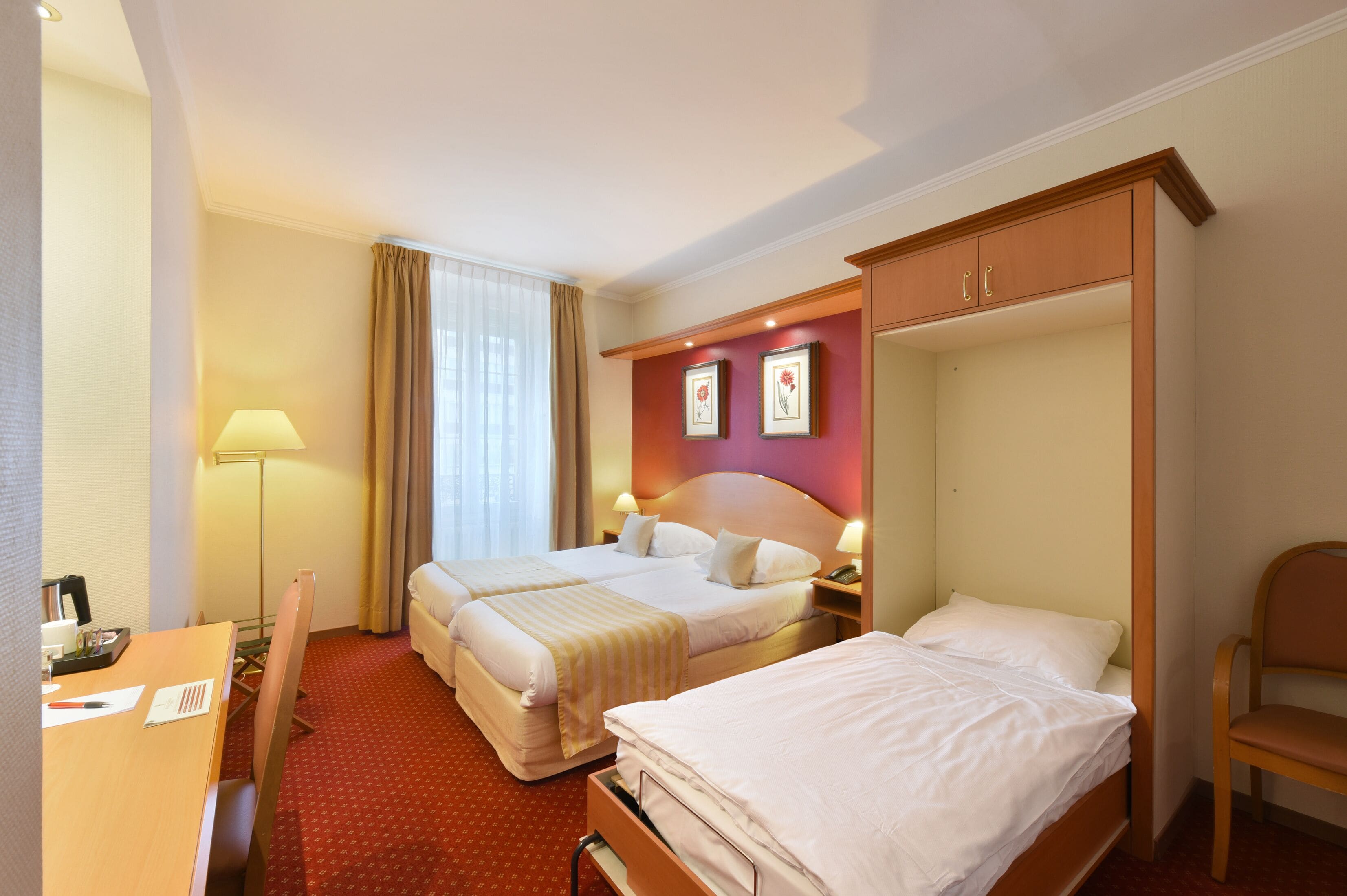 A hotel room with two beds, a desk, a chair, a lamp, and a wardrobe.