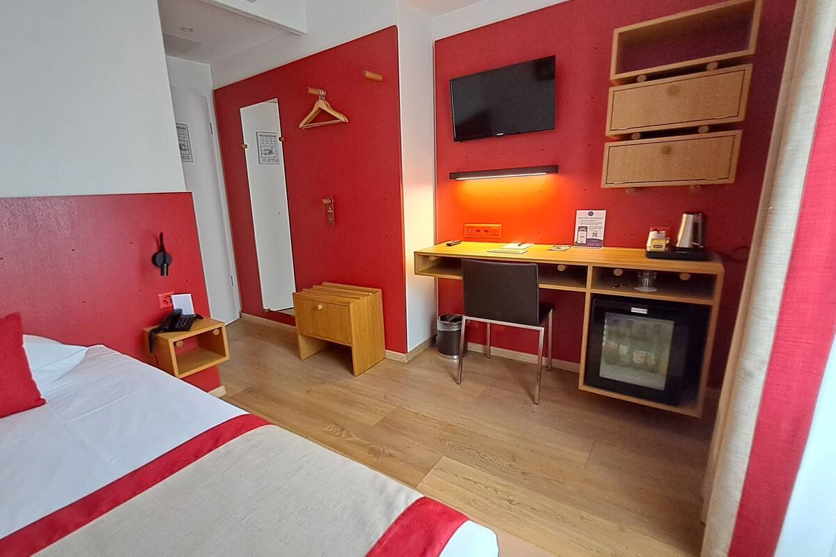 A compact hotel room with a bed, a desk, a chair, a small cabinet, a television, and a refrigerator.