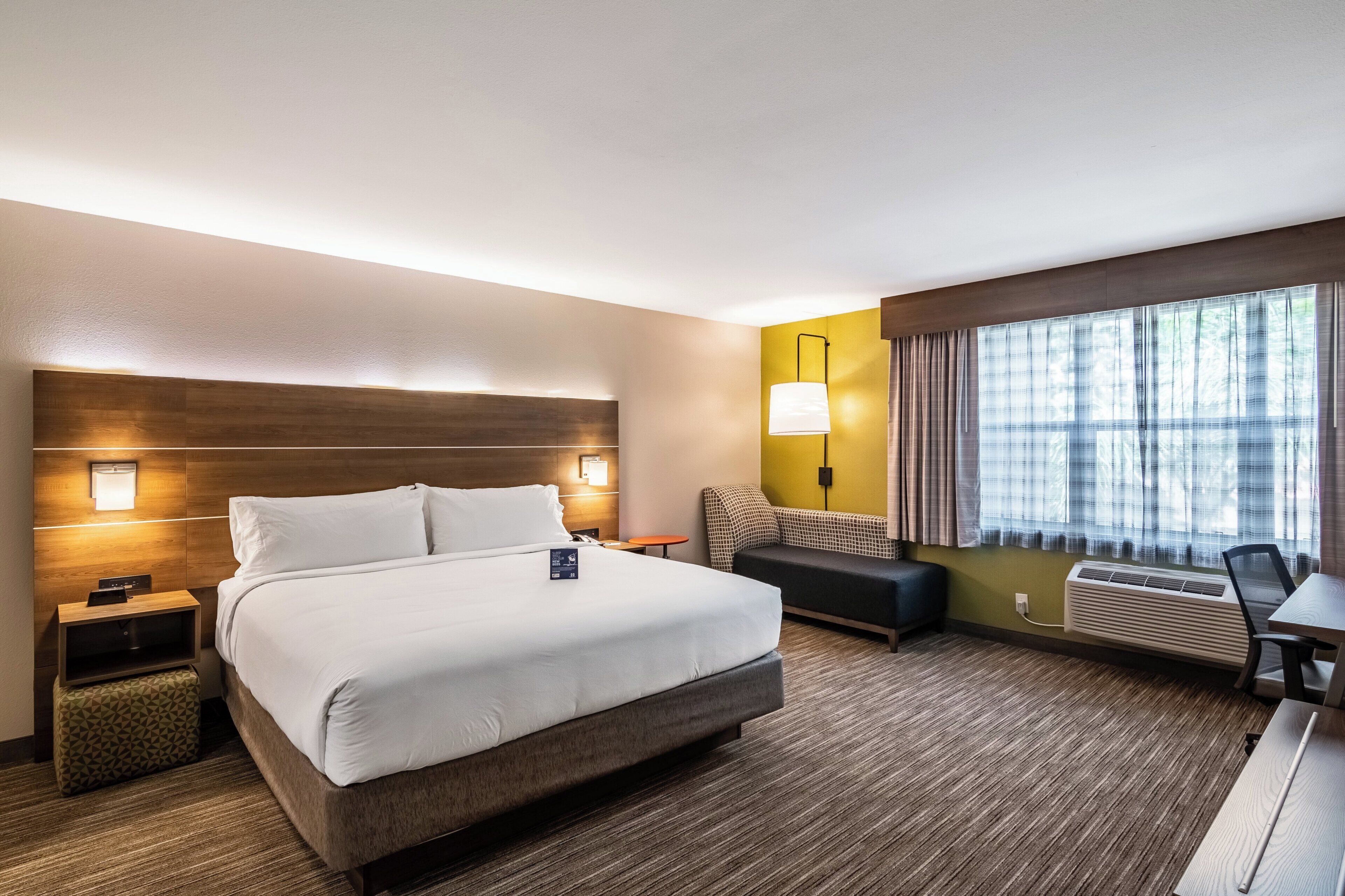 Ein Hotelzimmer mit einem großen Bett, Nachttischen, einem Schreibtisch, einem Sessel, einer Couch, einer Lampe und einem Vorhangfenster.