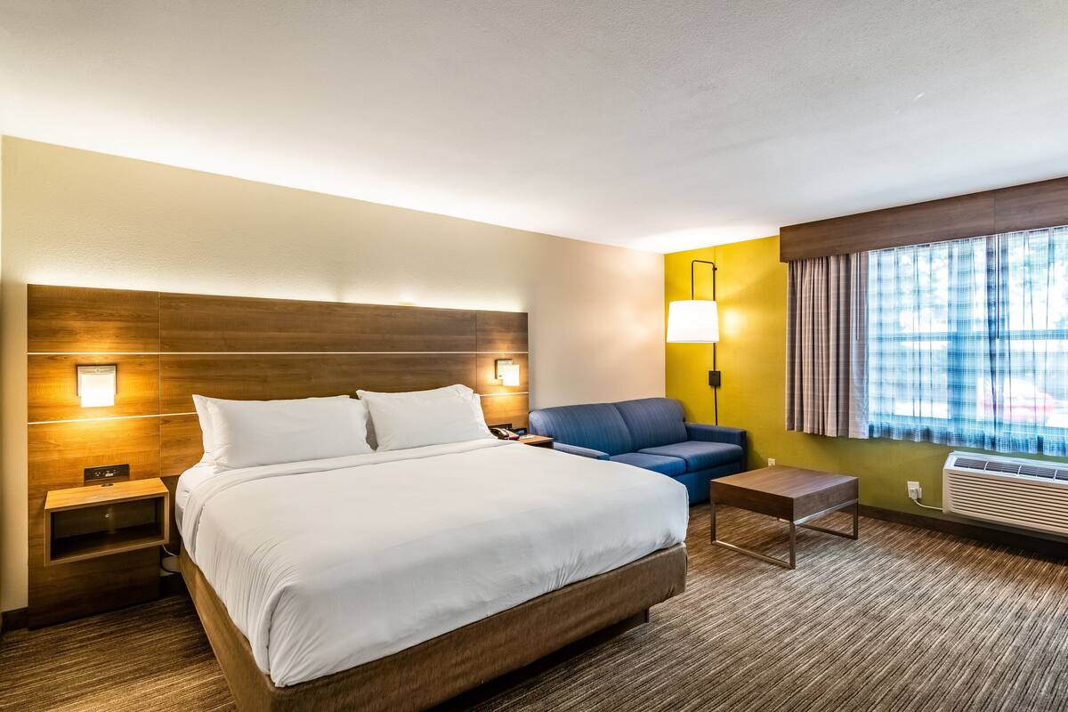A hotel room with a large bed, a bedside table, a nightstand, a sofa, a lamp, and a window with curtains.