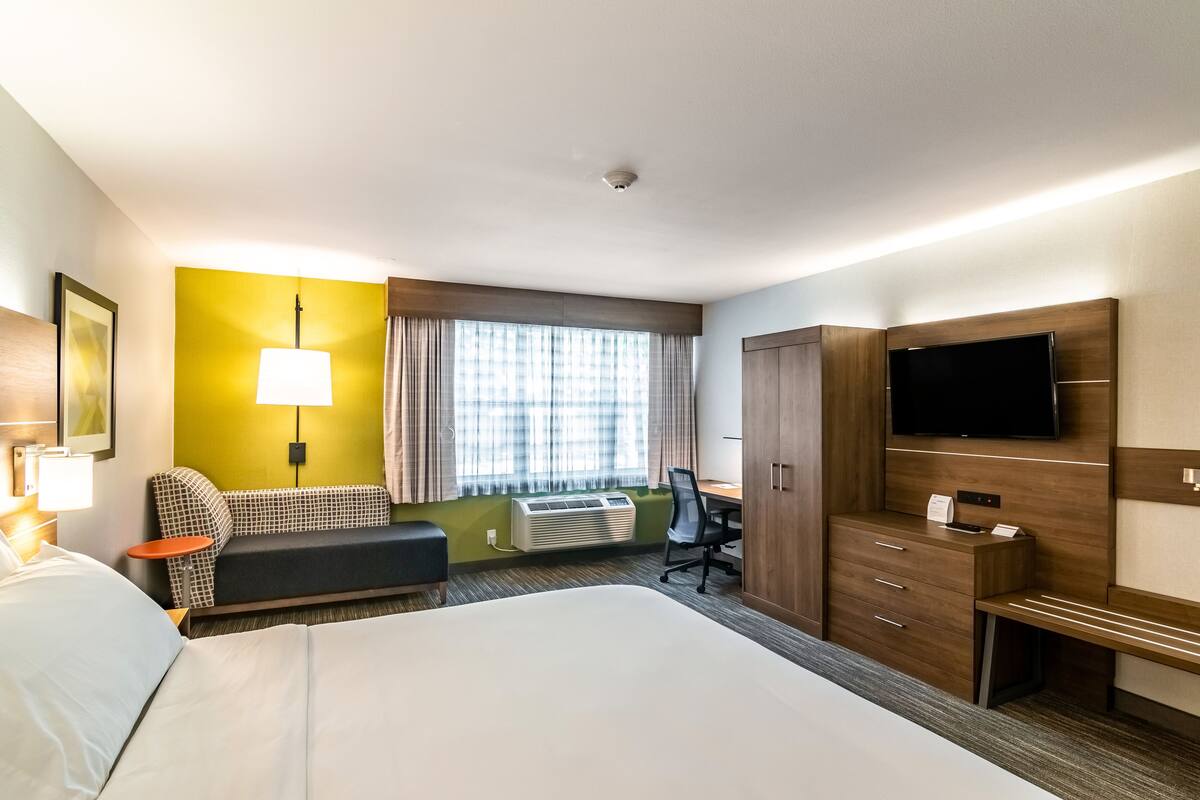 A hotel room with a large bed, a desk with a chair, a TV mounted on the wall, and a window with curtains.