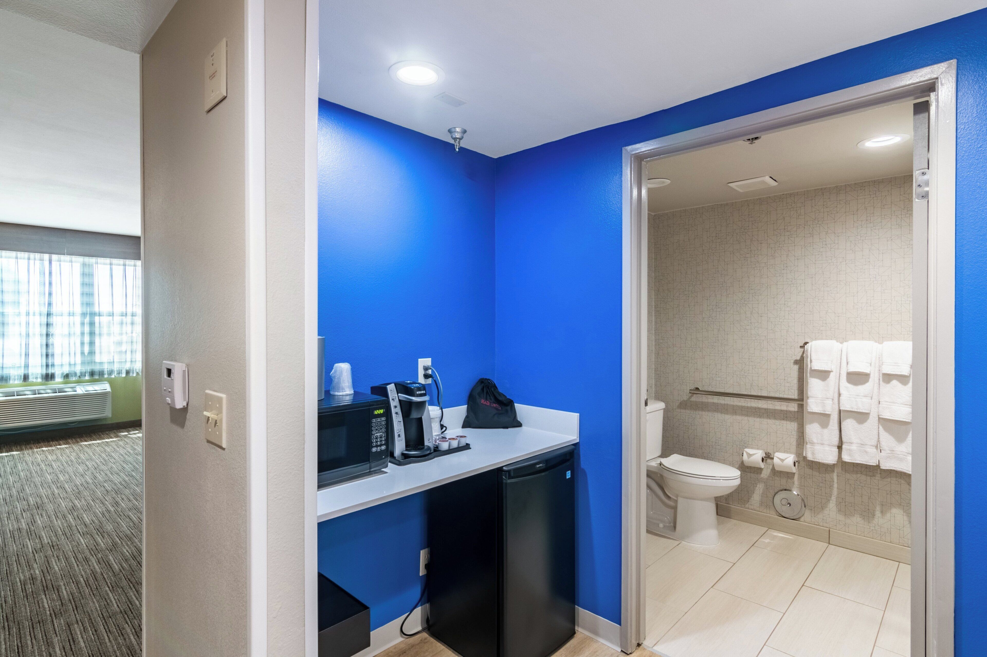 Un bany d'hotel amb una paret blava, un vàter, un lavabo amb una màquina de cafè i un gresol.