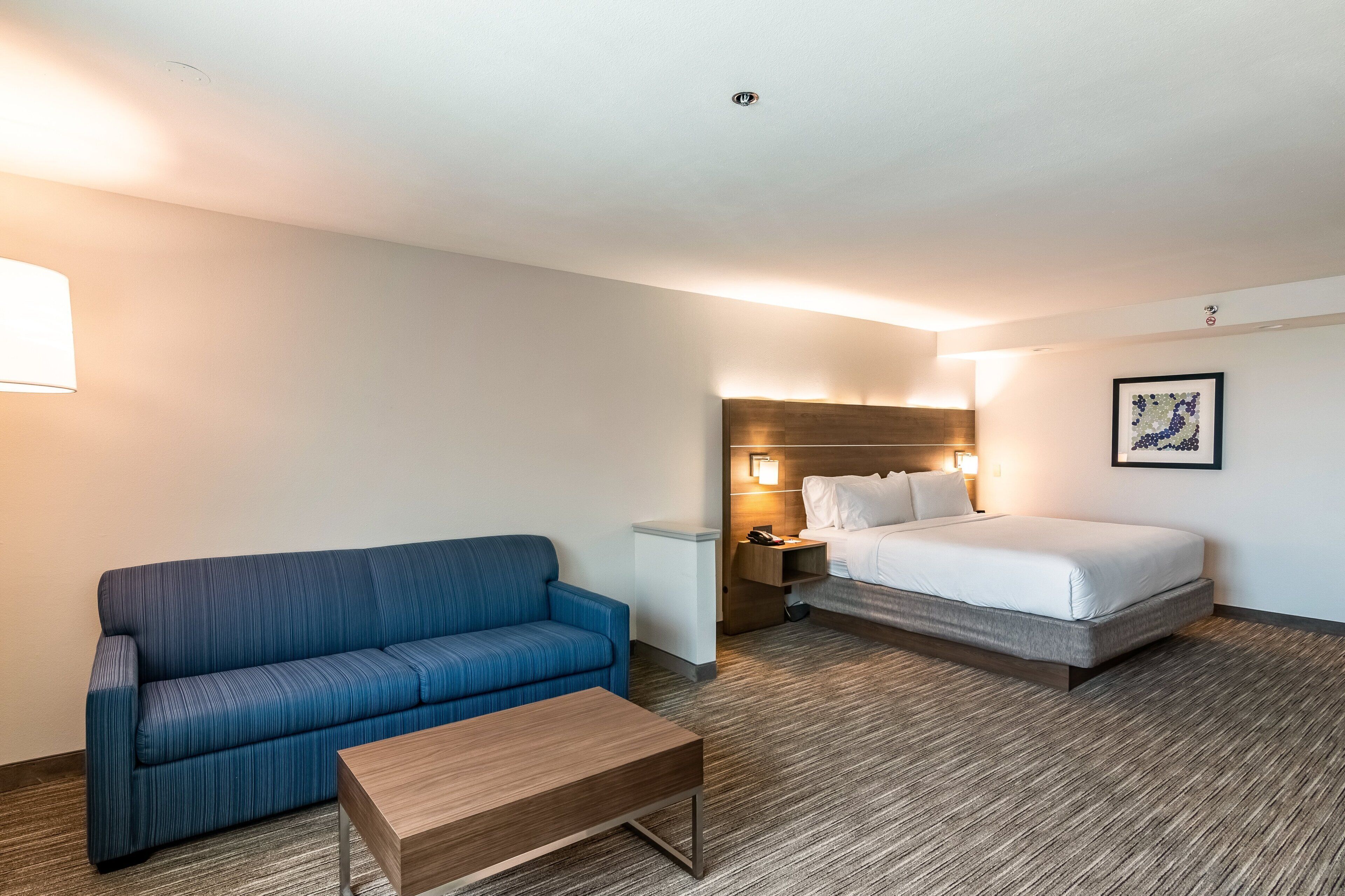 Une chambre d’hôtel avec un canapé bleu, une table basse en bois, un lit avec une literie blanche, une table de chevet et une lampe fixée au mur.