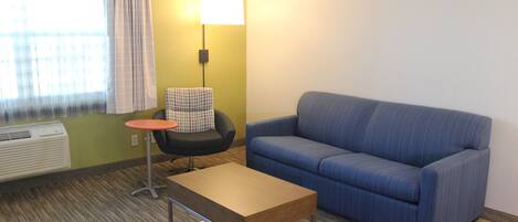 A room with a blue sofa, a small round table, a chair, a lamp, and a window with curtains.