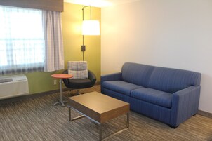 Una habitación con un sofá azul, una mesa redonda pequeña, una silla, una lámpara y una ventana con cortinas.