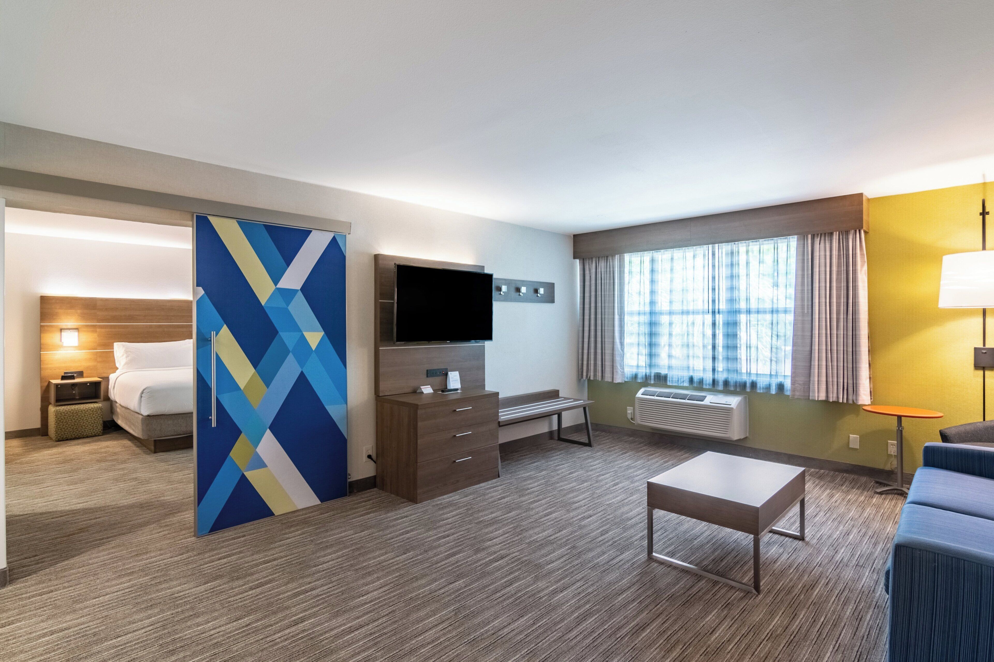 A hotel room with a bed, a desk, a television, a window with curtains, and a small table.