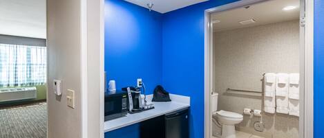 Un baño pequeño con una pared azul de acento, un lavamanos con cafetera, un refrigerador negro y un inodoro con toallero.