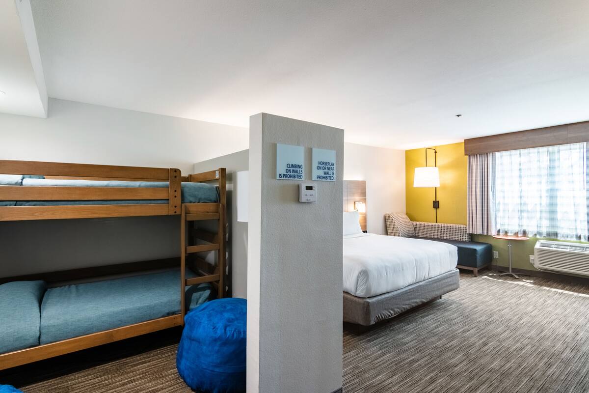 A hotel room with a bunk bed, a single bed, a desk, and a lamp. There are signs prohibiting climbing on walls and horseplay.