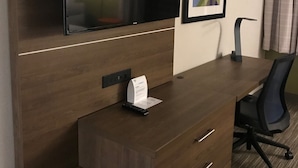 A hotel room with a wooden desk, a flat-screen TV mounted above it, a framed abstract artwork on the wall, a black office chair, and a small table with a remote control.