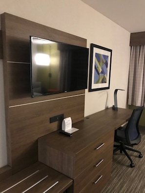 A hotel room with a wooden desk, a flat-screen TV mounted above it, a framed abstract artwork on the wall, a black office chair, and a small table with a remote control.