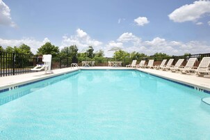 A rectangular swimming pool with clear blue water, surrounded by a black fence. There are lounge chairs and umbrellas along the poolside.
