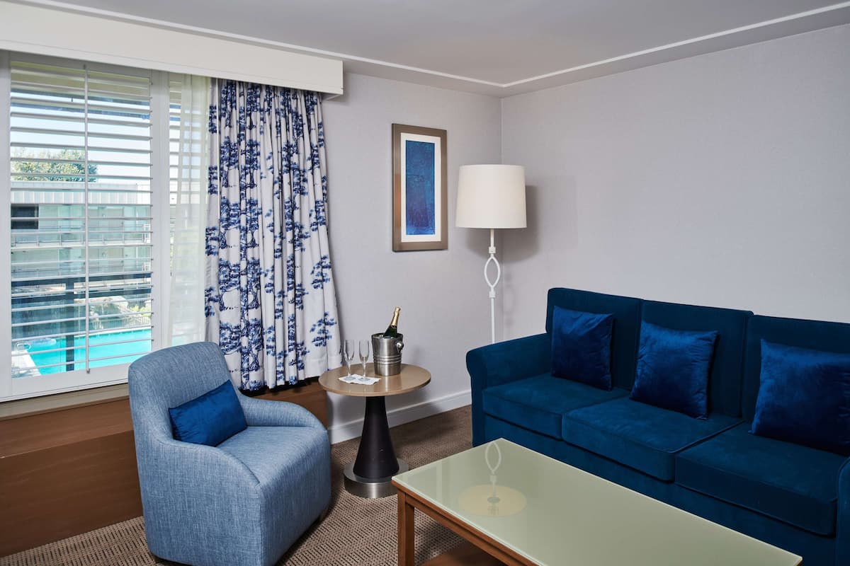 A modern living room with a blue sofa, a blue armchair, a glass coffee table, a small round table with a lamp, and a window with blinds.