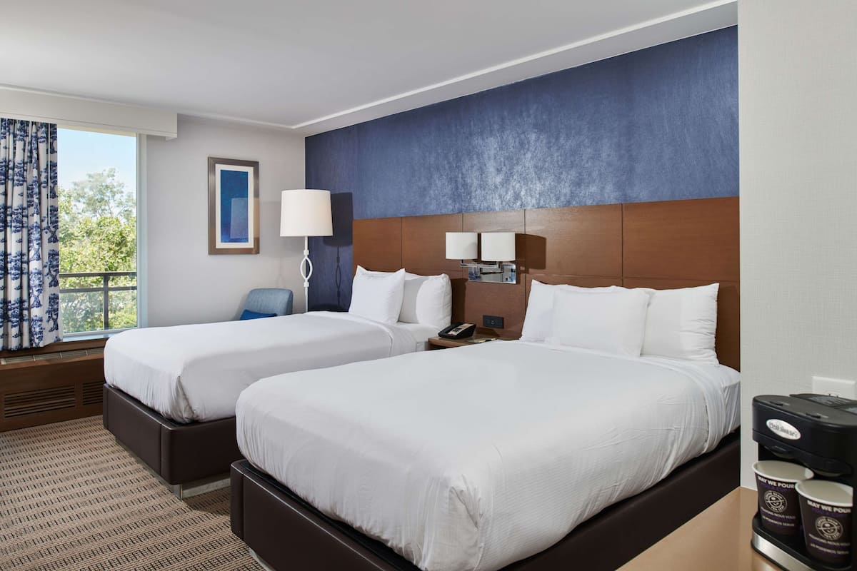 A hotel room with two beds, a large window with curtains, a wall-mounted lamp, and a coffee maker.