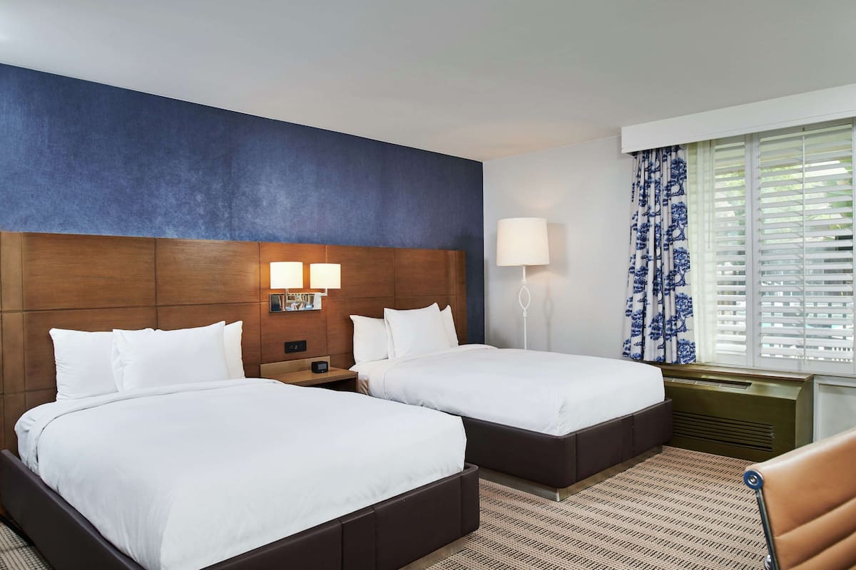 A hotel room with two beds, a wooden headboard, a nightstand with a lamp, a window with blinds, and a patterned rug.