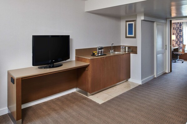 A compact hotel room with a kitchenette, a flat-screen TV, a small coffee maker, and a sink.