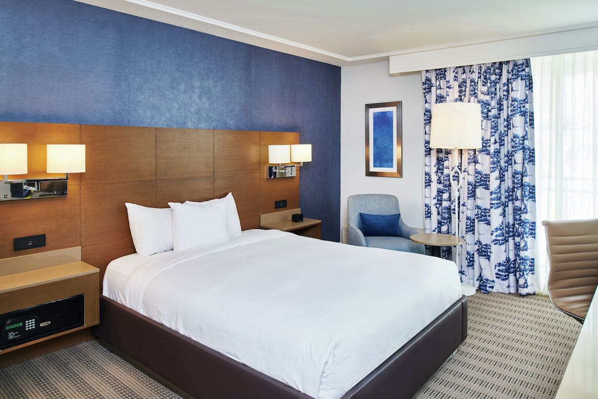 A hotel room with a large bed, bedside tables with lamps, a chair, a small table, and a window with curtains.