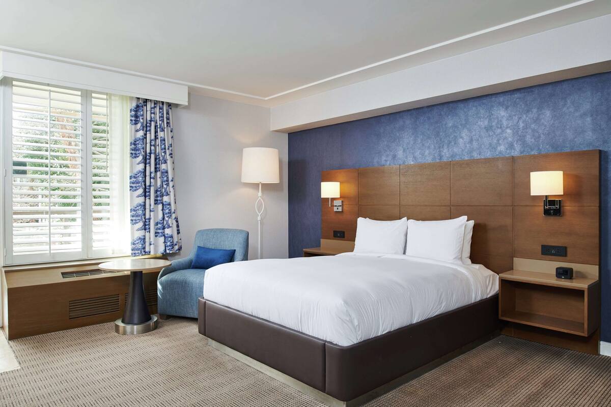 A modern hotel room with a large bed, two bedside tables with lamps, a chair, a window with blinds, and a built-in heater.