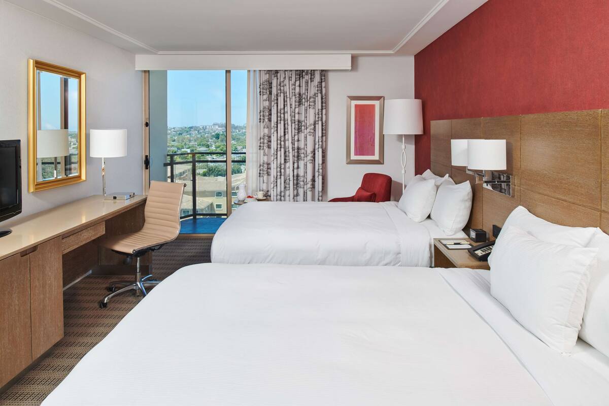 A hotel room with two beds, a desk, a chair, a TV, and a view of the city.