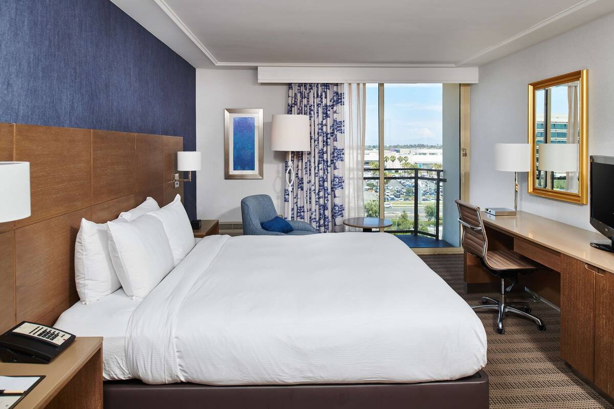 A hotel room with a large bed, a desk, a chair, a TV, and a view of the city.