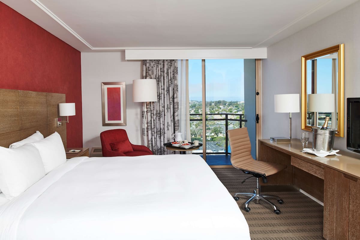 A hotel room with a large bed, a desk, a red chair, a small table with a pitcher and cups, and a large window with a view.