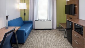 A compact hotel room with a blue sofa, a small desk, a television, a mini-fridge, and a window with curtains. - Holiday Inn Express Atlanta Airport - College Park by IHG (College Park)