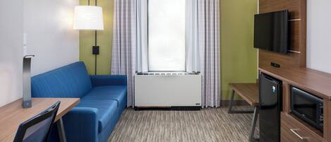 Habitación de hotel compacta con sofá azul, escritorio pequeño, televisor, mini refrigerador y ventana con cortinas.