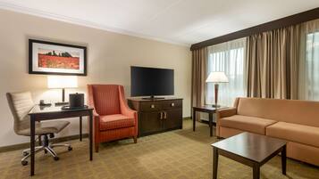A hotel room with a desk, chair, sofa, TV, and a framed picture on the wall.