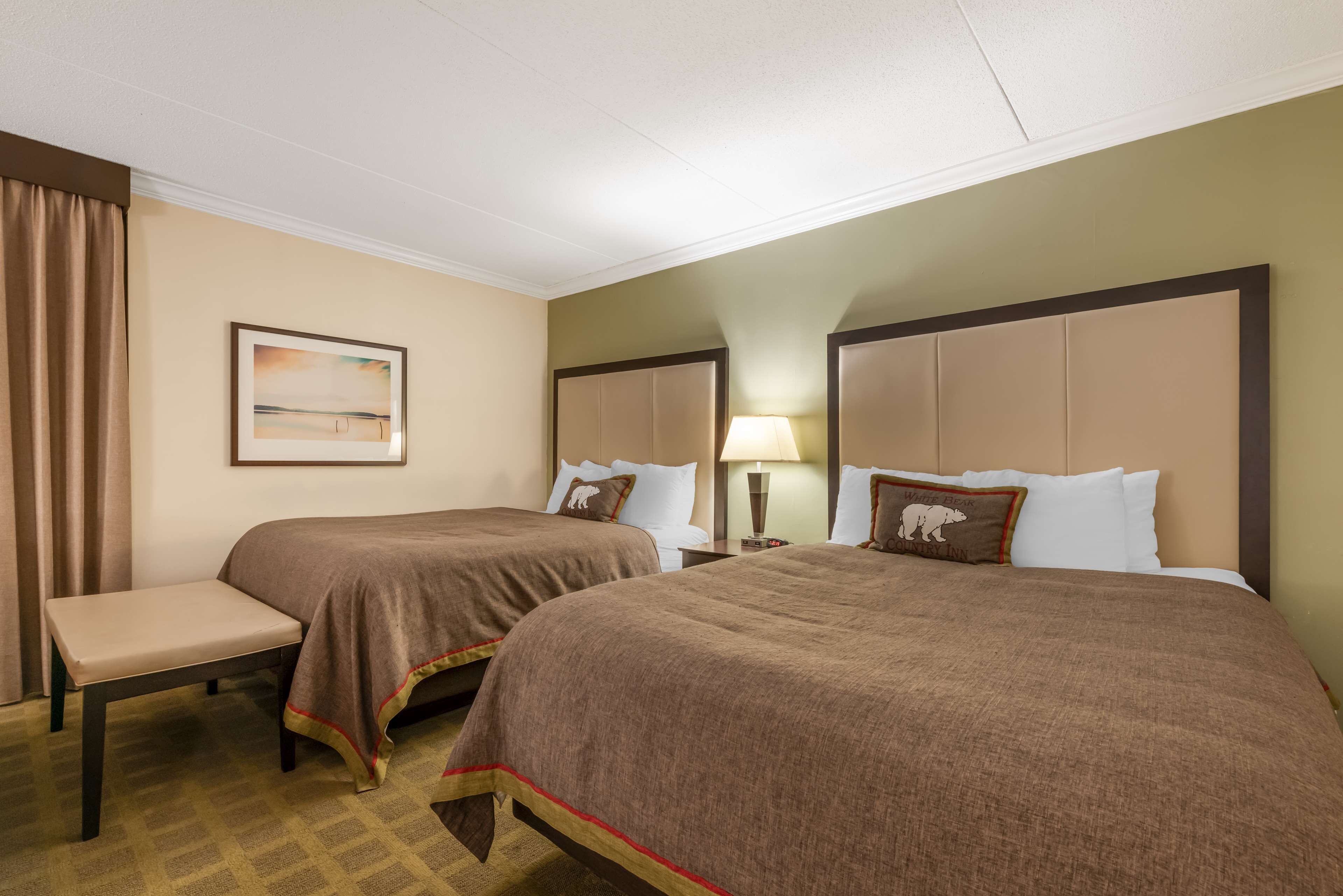 A hotel room with two beds, a bench, a painting, and a lamp.