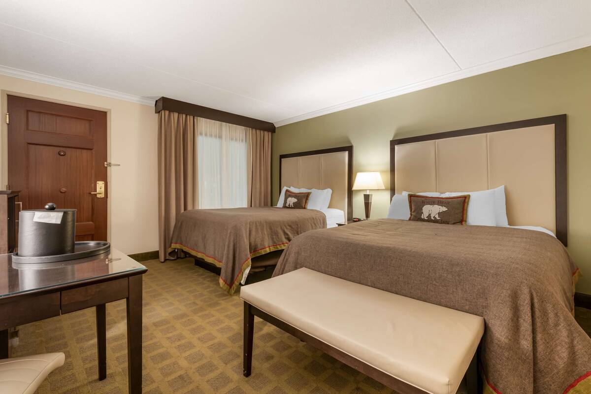 A hotel room with two beds, a bench, a desk with a kettle, and a door.