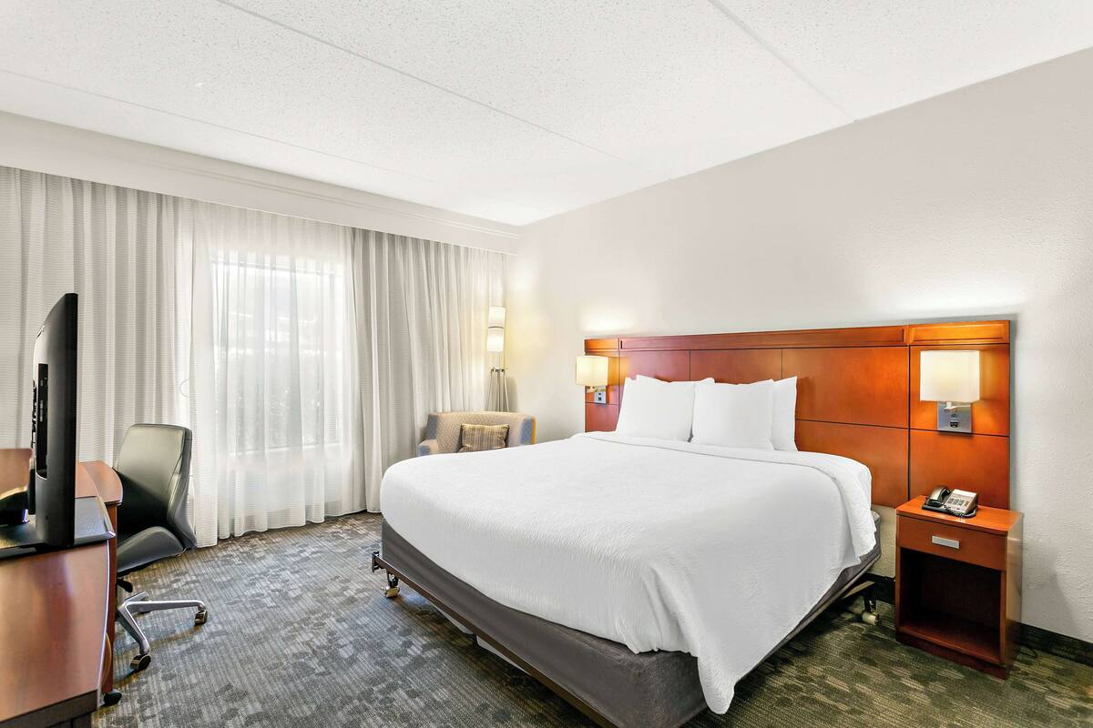 Ein Hotelzimmer mit einem großen Bett, einem Schreibtisch mit Computer, einem Stuhl, einer Lampe und einem Telefon auf einem Nachttisch.
